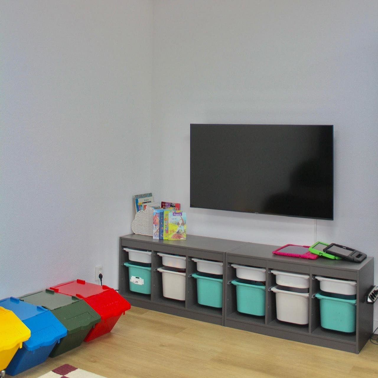 Een lichte ruimte met een kast vol opbergbakken, boeken en tablets onder een televisie.