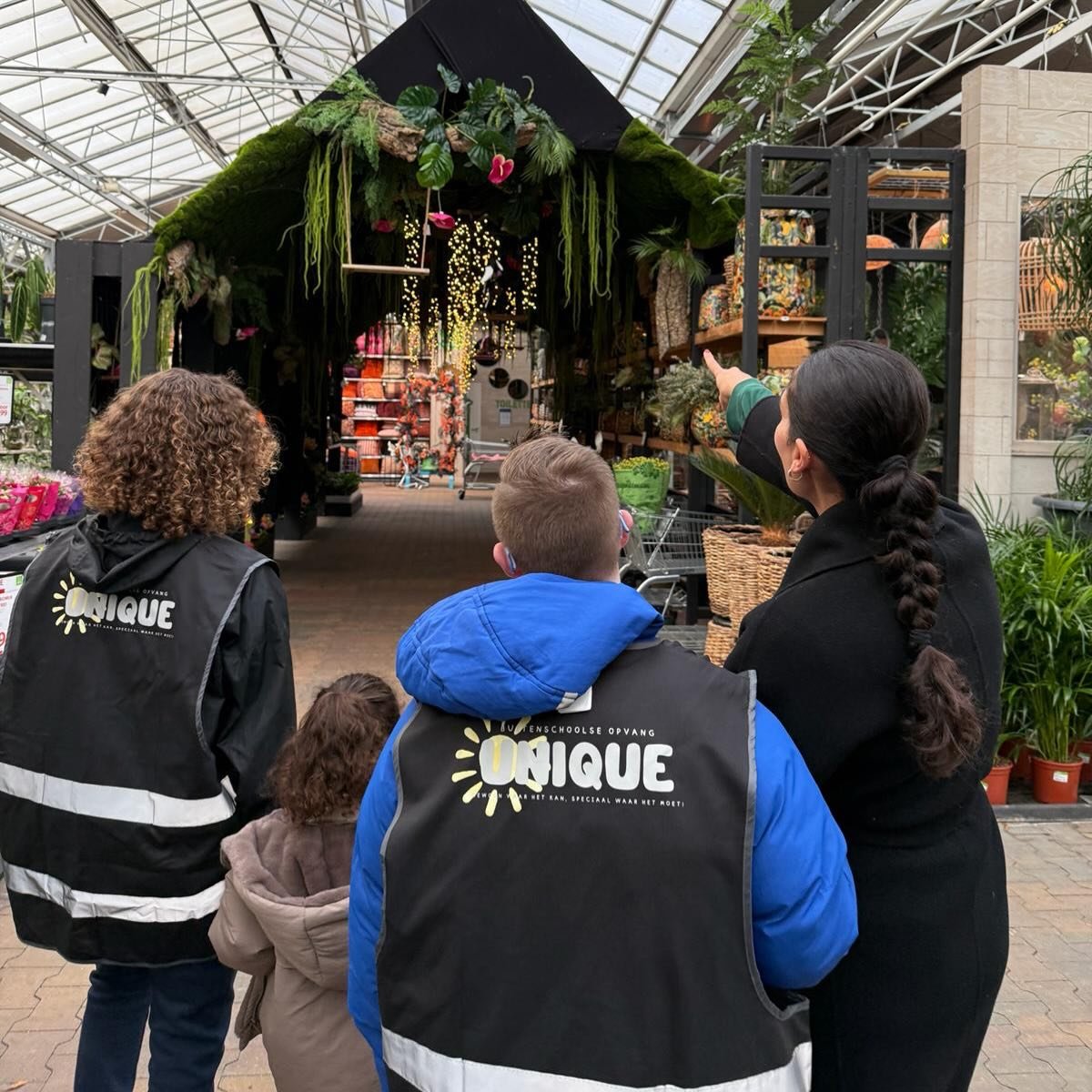 Een begeleider van Unique wijst iets aan tijdens een uitje naar het tuincentrum; drie deelnemers in Unique-hesjes kijken mee.